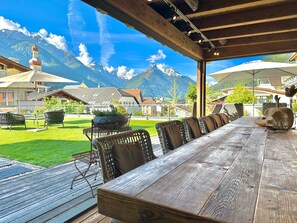 A wooden deck with a long table and chairs, overlooking a mountainous landscape with a church spire visible in the background. - Aparthotel Krösbacher (Fulpmes)