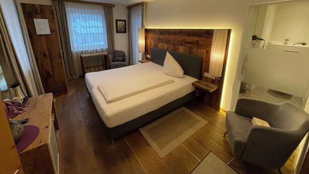A hotel room with a large bed, wooden headboard, a desk with a chair, a small table, and a mirror. - Aparthotel Krösbacher (Fulpmes)