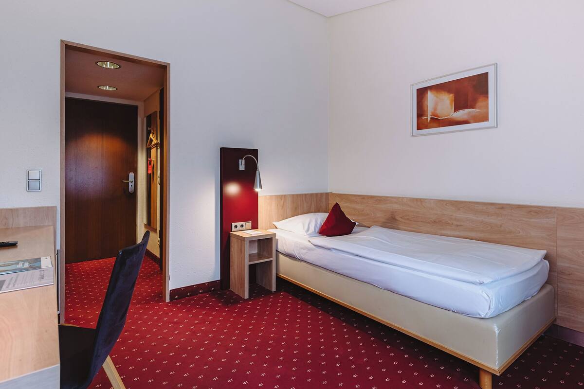 A hotel room with a single bed, a desk with a chair, a small bedside table, a wall-mounted lamp, and a red carpet.