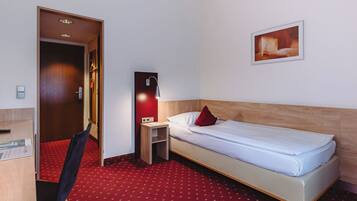 Habitación de hotel con una cama individual, un escritorio con silla, una mesita de noche pequeña, una lámpara de pared y una alfombra roja.