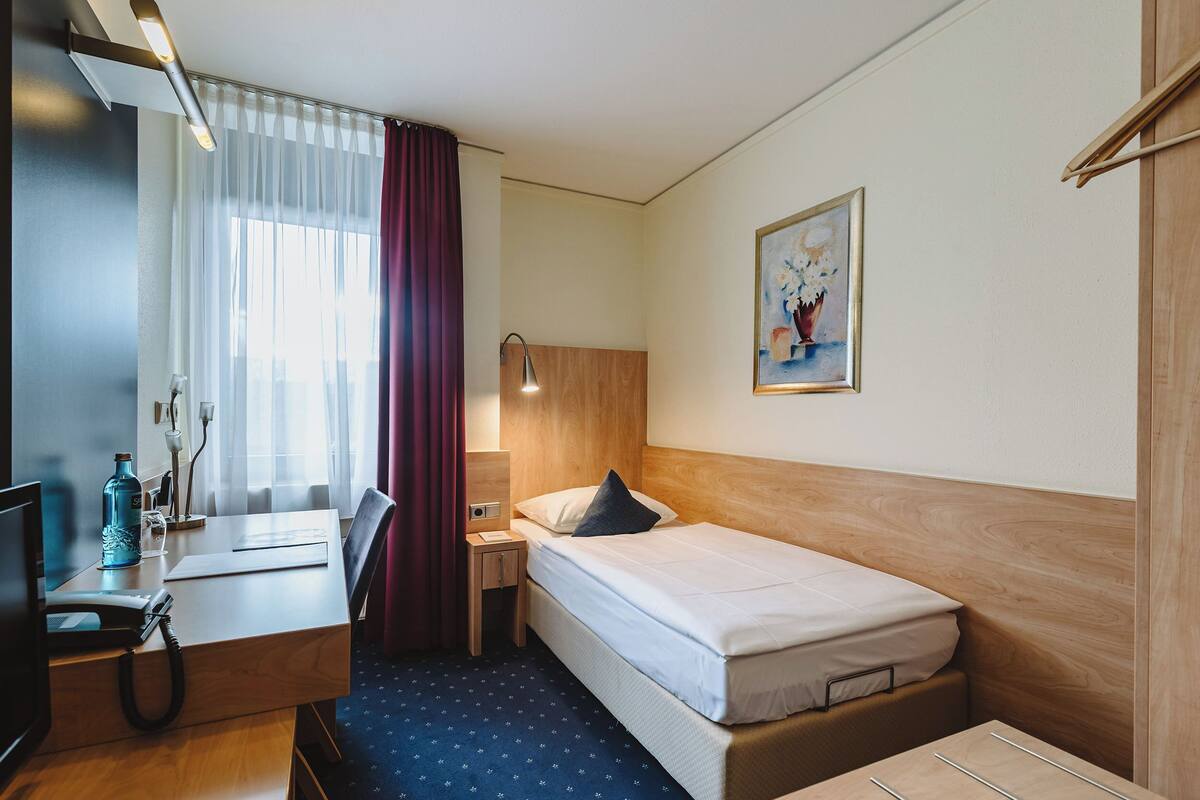 A hotel room with a bed, desk, chair, lamp, and a painting on the wall.