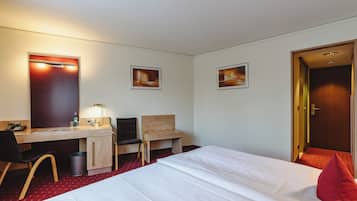 A hotel room with a bed, desk, chair, and a small table with a lamp.