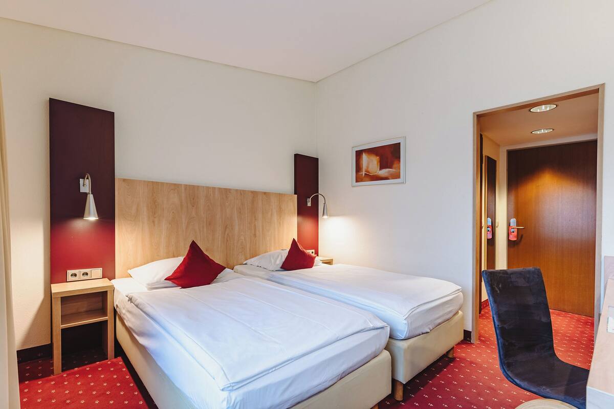 A hotel room with two beds, a small table, a chair, and a door.