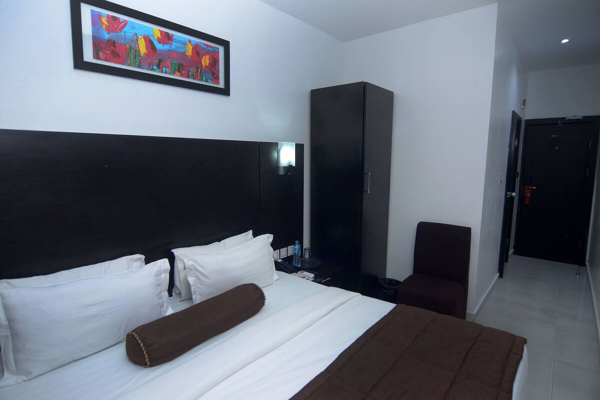 A hotel room with a large bed, a wardrobe, a chair, a desk, and a painting on the wall.