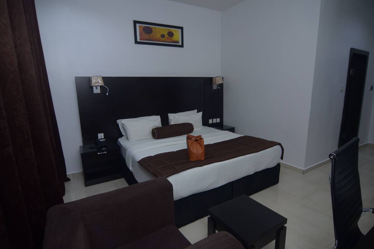 A hotel room with a bed, bedside tables, a chair, a small table, and a painting on the wall.