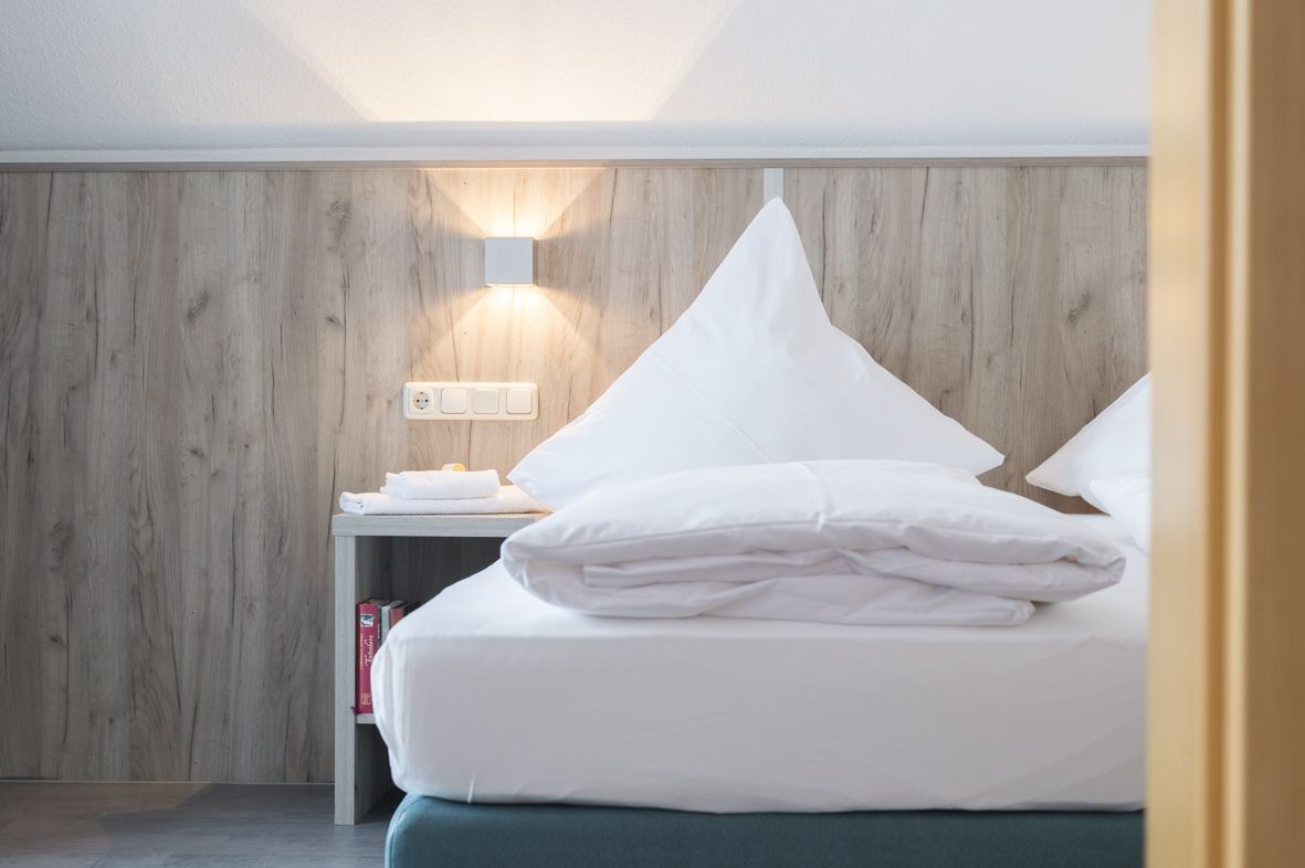 Ein ordentlich bezogenes Bett mit weißen Leinen und Kissen, ein Nachttisch mit Lampe sowie eine Holzvertäfelung an der Wand.