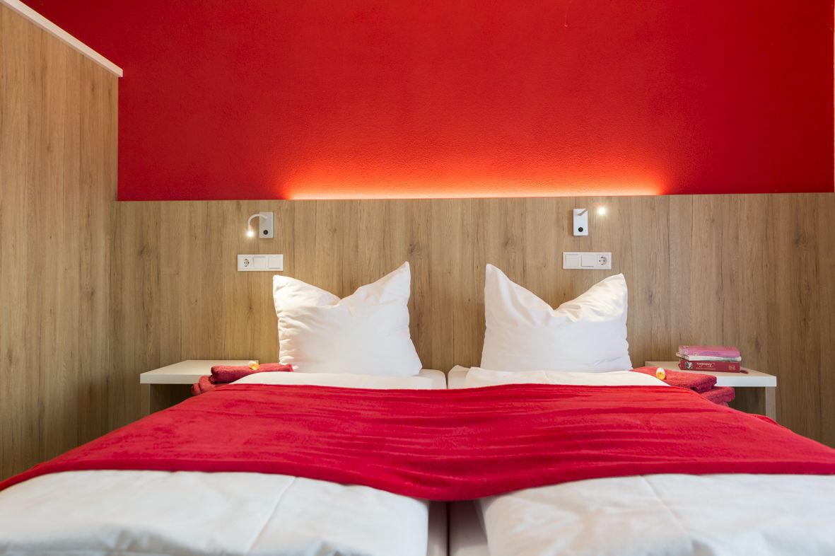 Ein Doppelbett mit weißer Bettwäsche und roter Decke, zwei Nachttische mit Lampen sowie eine Wand in roter Farbe.