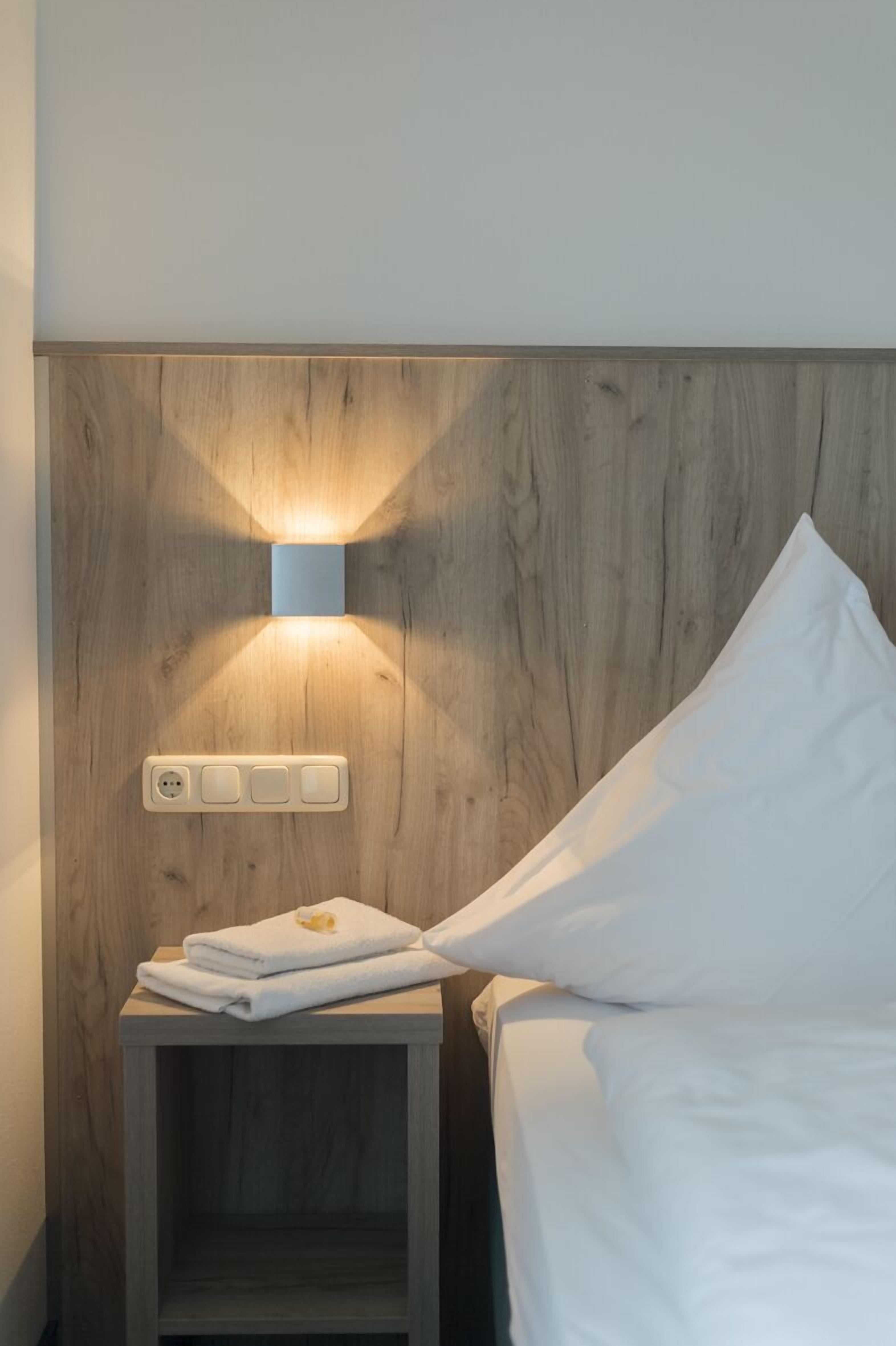 Una cama bien hecha con sábanas blancas, una mesita de noche de madera con una lámpara fijada a la pared y un enchufe.