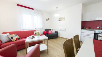 A modern living room with a red sofa, a grey sofa, a dining table, and a kitchen area with white cabinets and a stove.