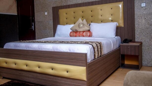 A neatly made bed with a tufted headboard, a bedside table with a phone and a book, and a small wooden cabinet. - Le Brigadier Hotel (Lekki)