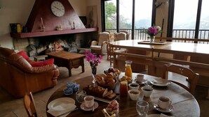A cozy living room with a fireplace, a dining area with breakfast spread, and a view of mountains through large windows. - San Larenzu (Aullène)