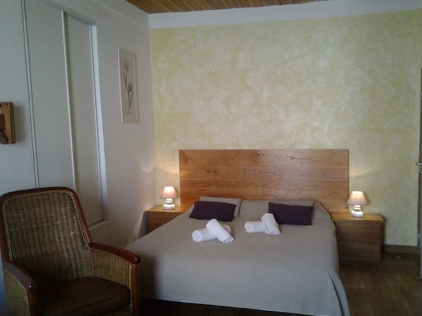 A bedroom with a wooden headboard, two bedside tables with lamps, a wicker chair, and two rolled towels on the bed. - San Larenzu (Aullène)