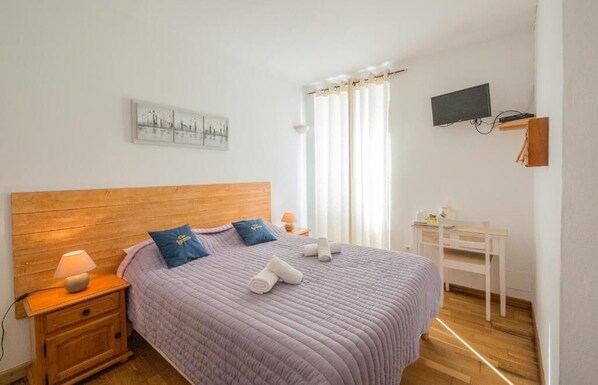 Un dormitorio con cabecera de madera, cama con colcha morada, dos almohadas, mesita de noche con lámpara, escritorio, televisor de pantalla plana montado en la pared y ventana con cortinas.