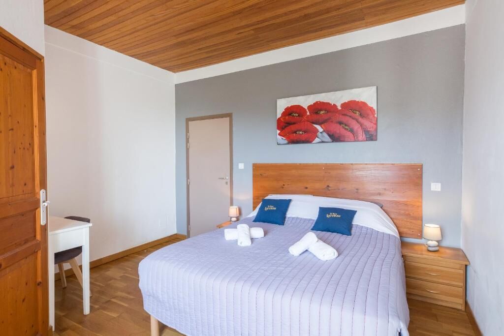 A hotel room with a wooden ceiling, a bed with a headboard, a painting of red flowers, and a desk with a chair.
