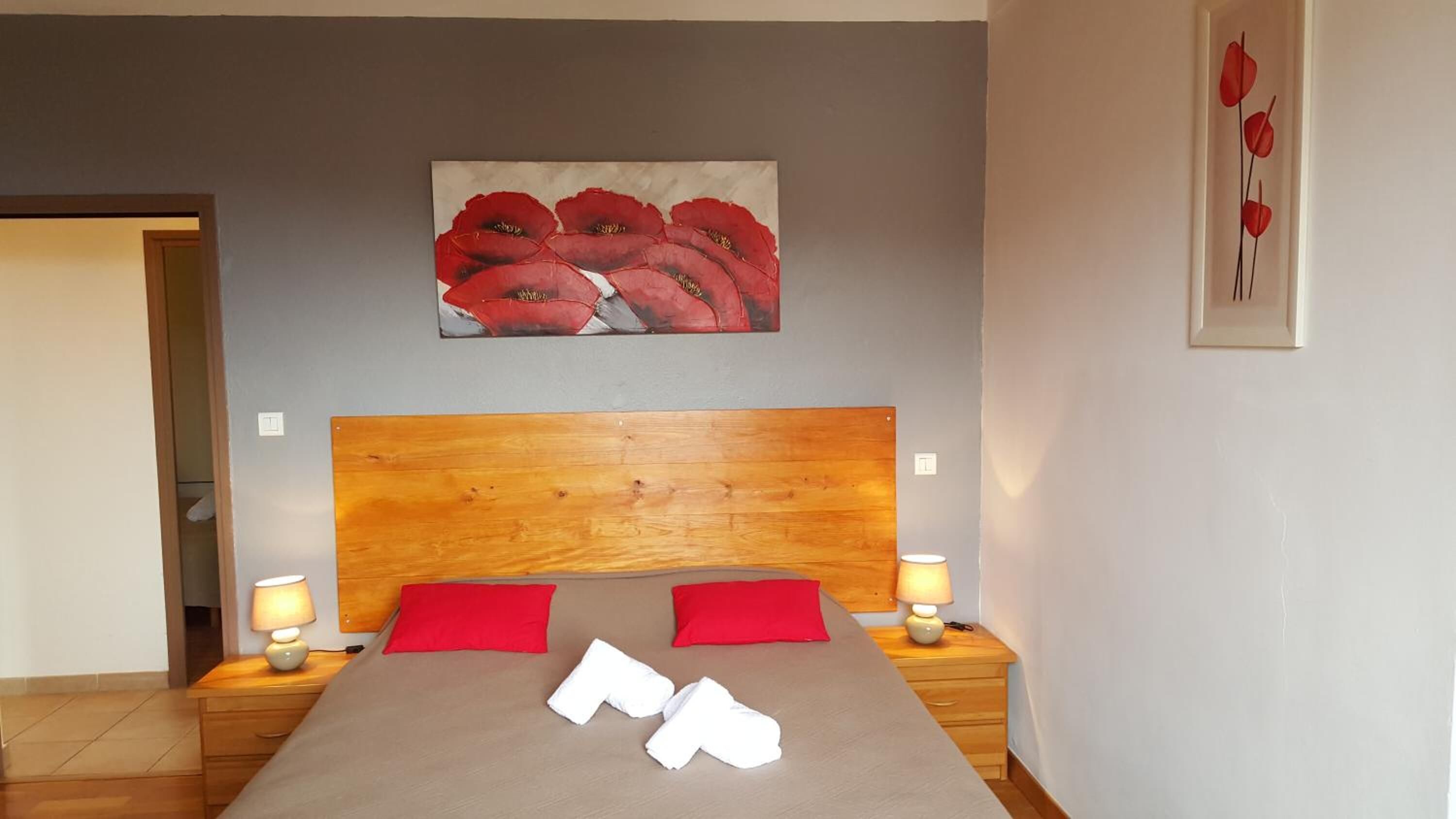 Un dormitorio con cabecera de madera, dos mesitas de noche con lámparas, una cama con almohadas rojas y blancas, y un cuadro de flores rojas en la pared.