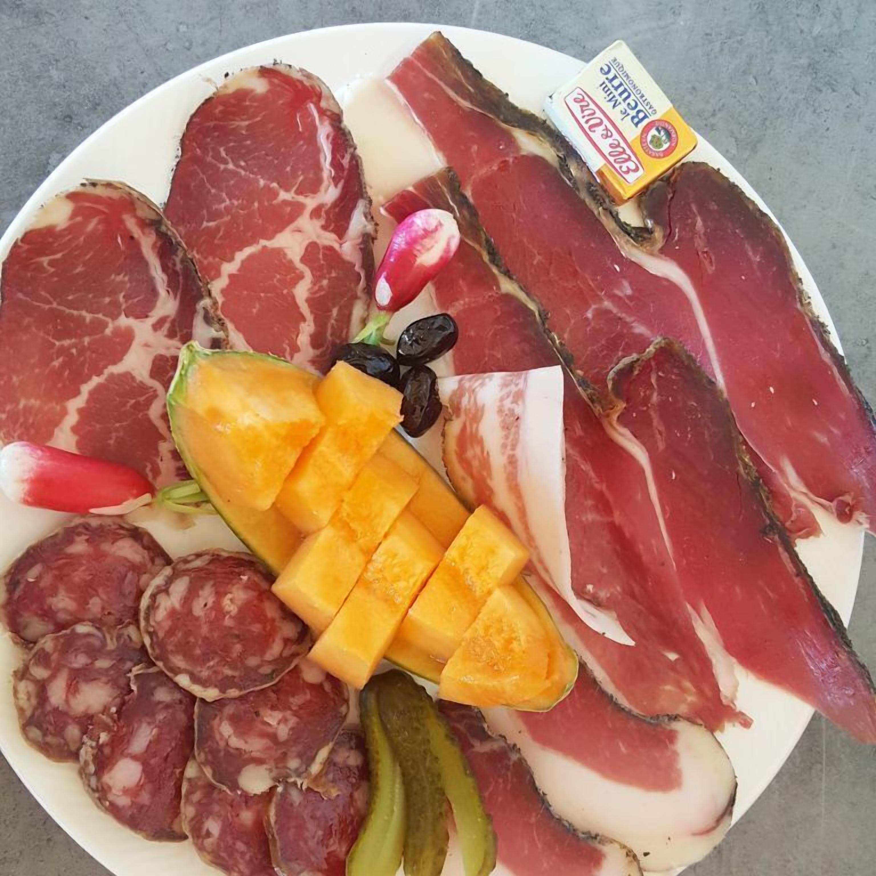 Een bord met diverse soorten vleeswaren, waaronder salami en parmaham, geserveerd met augurken, een mango en olijven.