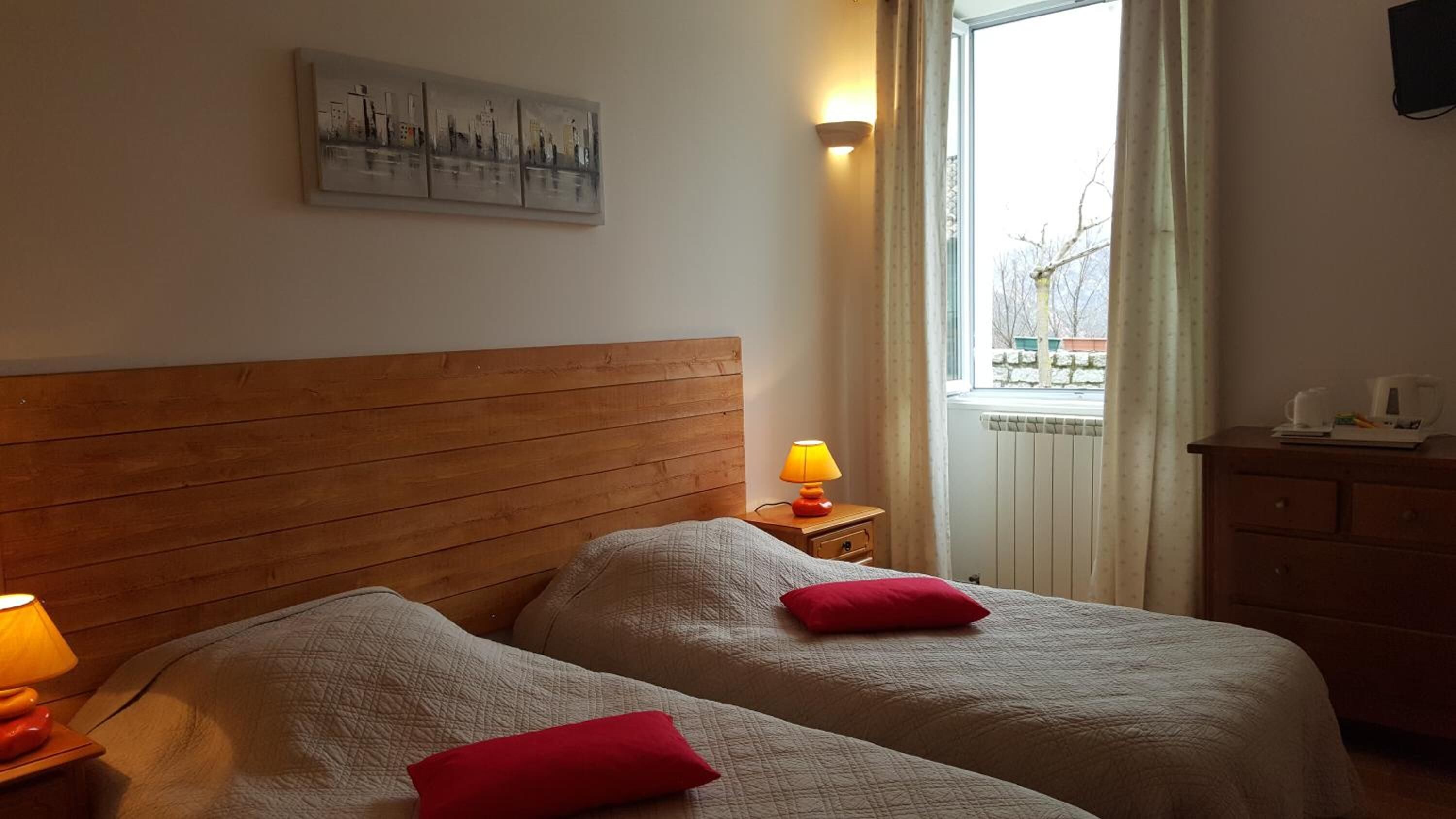 A room with two beds, a nightstand with a lamp, a dresser, a window with curtains, and a painting on the wall.