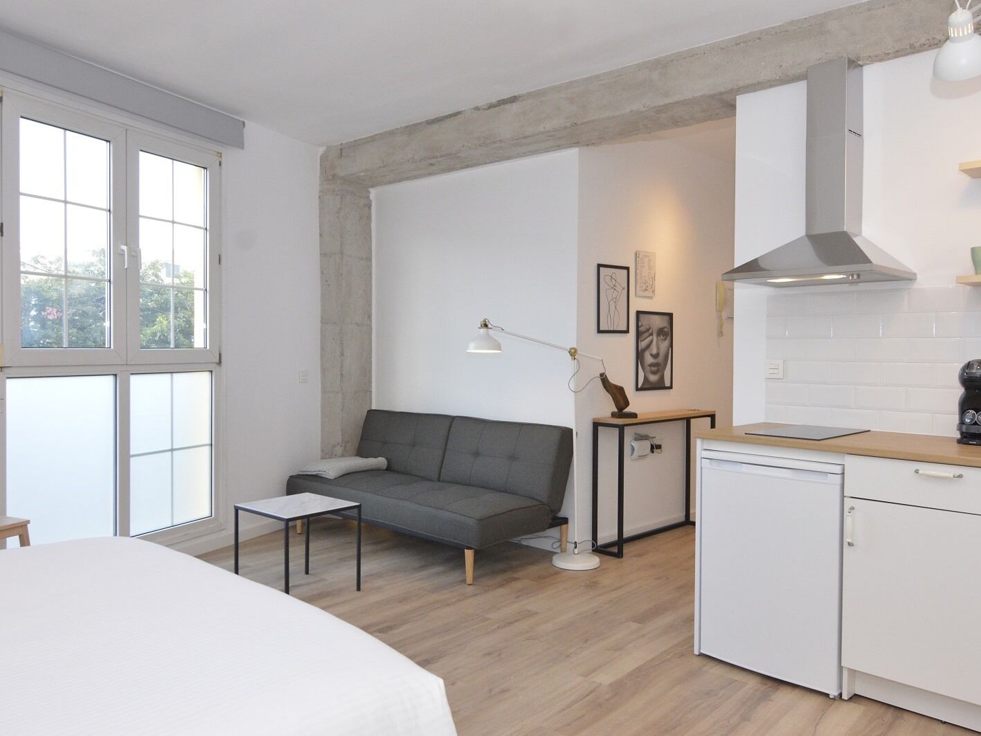 Una habitación moderna y minimalista con un sofá gris, una mesita pequeña, un armario blanco y un ventanal grande.