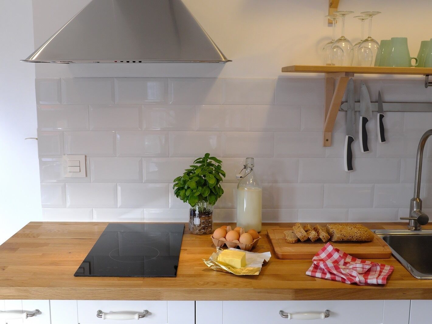 Una cocina con azulejos blancos, una placa de cocción por inducción negra, una planta, huevos, pan y una botella de leche.
