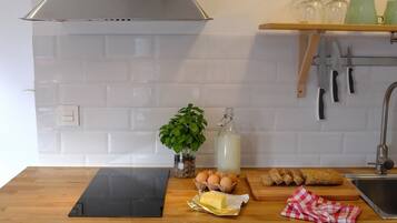 Una cocina con azulejos blancos, una placa de cocción por inducción negra, una planta, huevos, pan y una botella de leche.