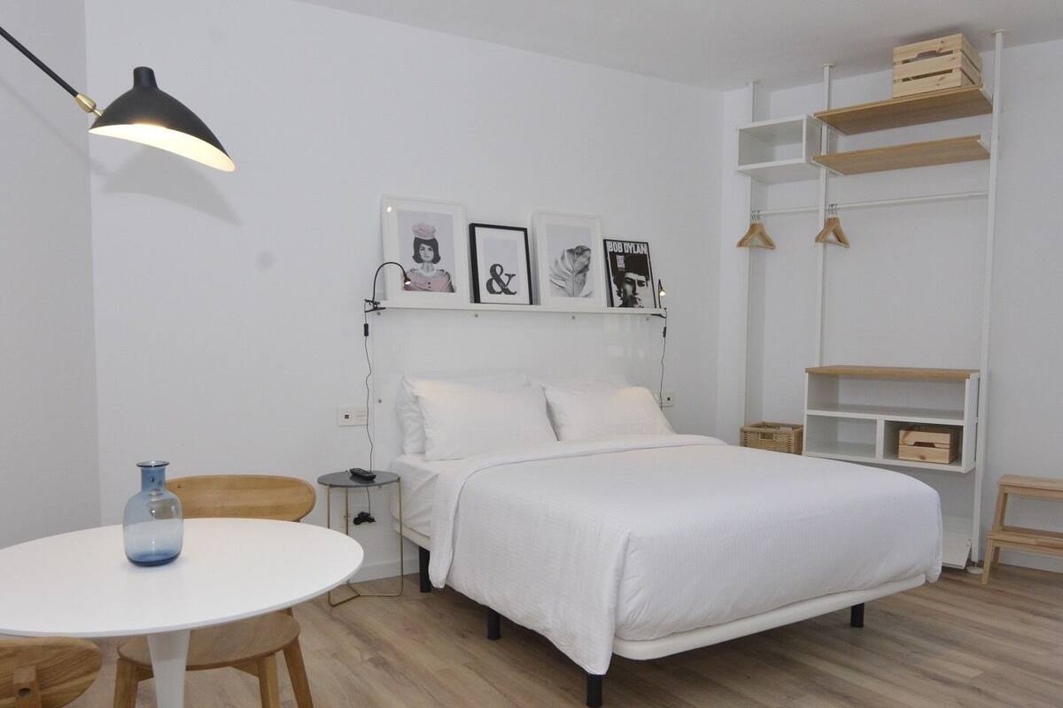 A minimalist bedroom with a bed, a bedside table, a lamp, and a small table with a vase.