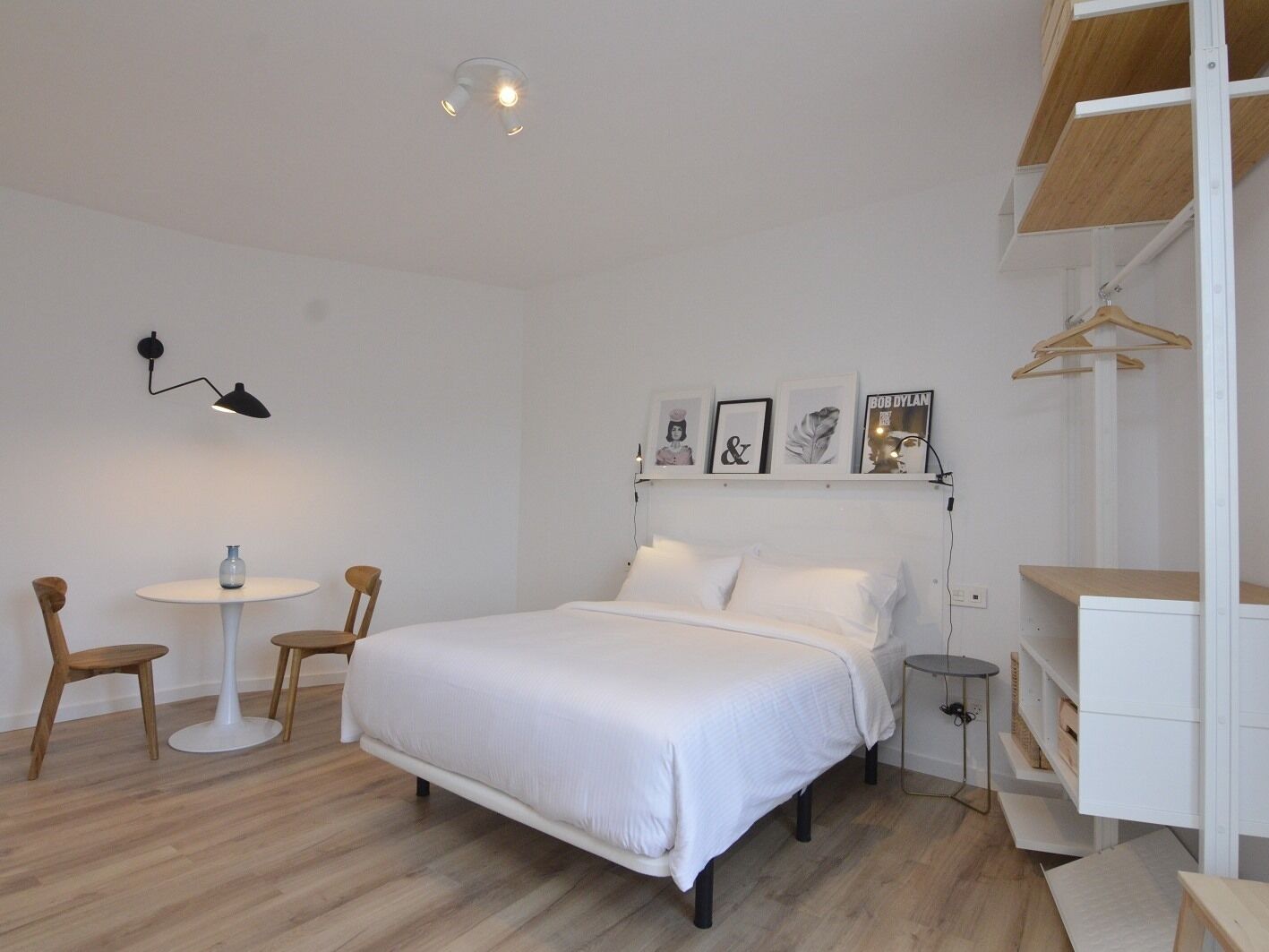 Un dormitorio con cama, mesita de noche, silla, lámpara colgante y estante con marcos de fotos.