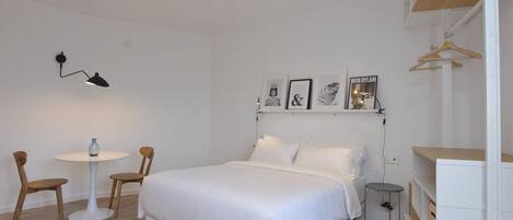 Un dormitorio con cama, mesita de noche, silla, lámpara colgante y estante con marcos de fotos.