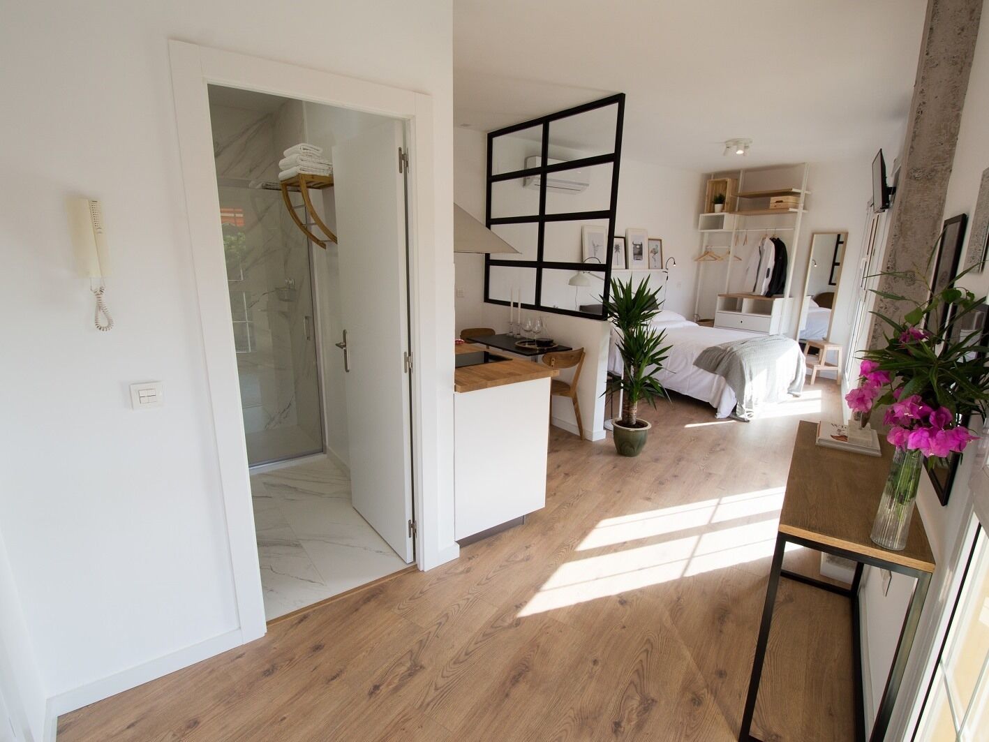 Moderni hotellihuone, jossa on puulattia, lasinen suihkukaappi, työpöytä, sänky ja ruukussa oleva kasvi.