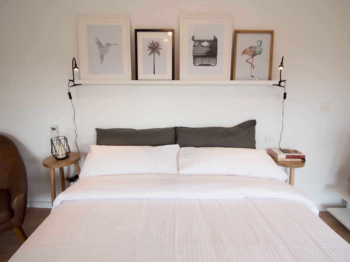 A bedroom with a large bed, two bedside tables, a chair, and a shelf with framed pictures.
