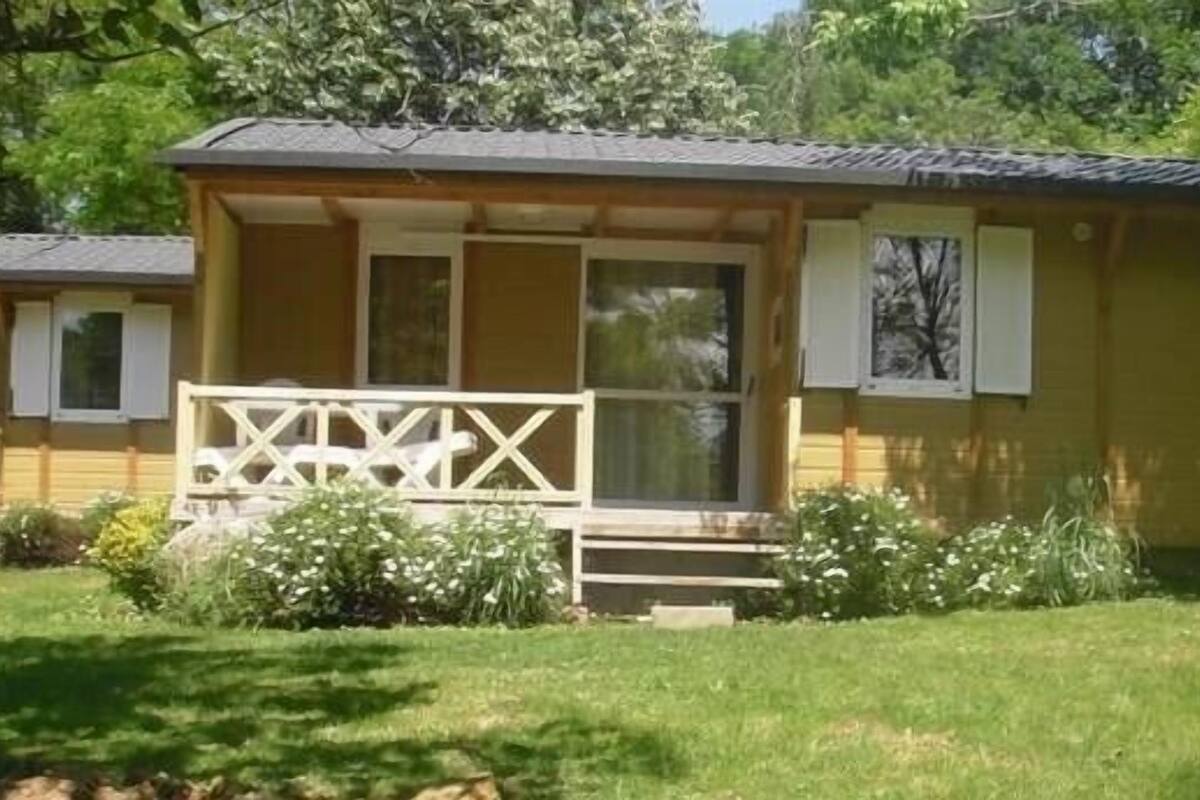 Une petite cabane en bois d’un seul étage, dotée d’un porche et entourée d’une cour verdoyante et d’arbres.