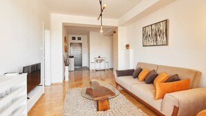 Uma sala de estar moderna com uma mesa de centro de vidro, um sofá bege com almofadas laranja, uma televisão de tela plana e uma área de jantar ao fundo.