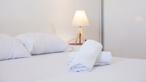 Cama bem arrumada com almofadas brancas e uma toalha enrolada, uma luminária de mesa e uma parede branca.
