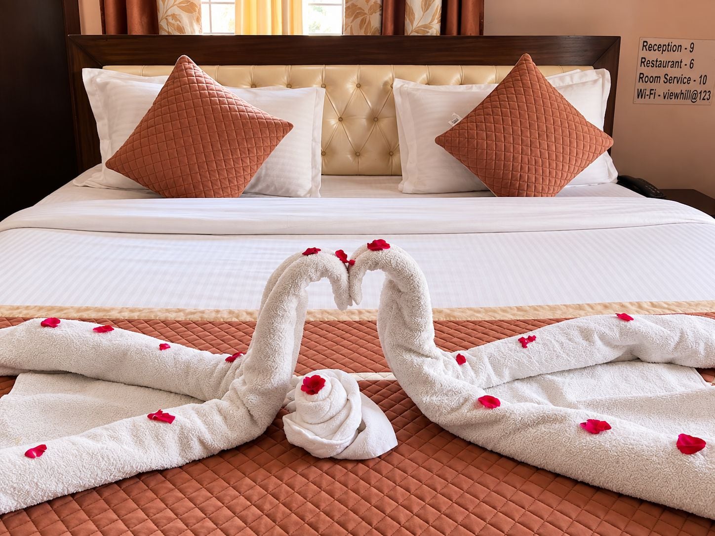 A hotel room with a bed, two pillows, a quilted bedspread, and a towel arrangement forming a heart shape.