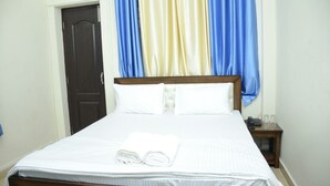 A neatly made bed with white linens and a folded towel, a wooden headboard, a small bedside table with a phone and glasses, and a door with a dark frame. - Hill View Homes (Nainital)