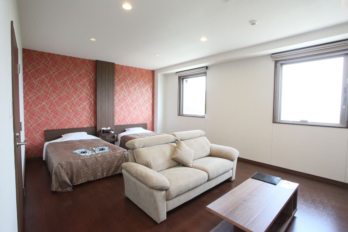 A hotel room with a bed, a sofa, a small table, and two windows.