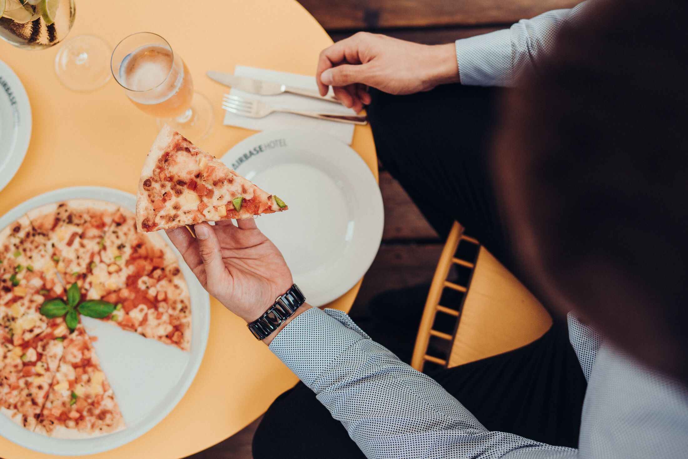 Une personne tenant une part de pizza à une table, devant une assiette de pizza, un verre d’eau, une fourchette et un couteau.