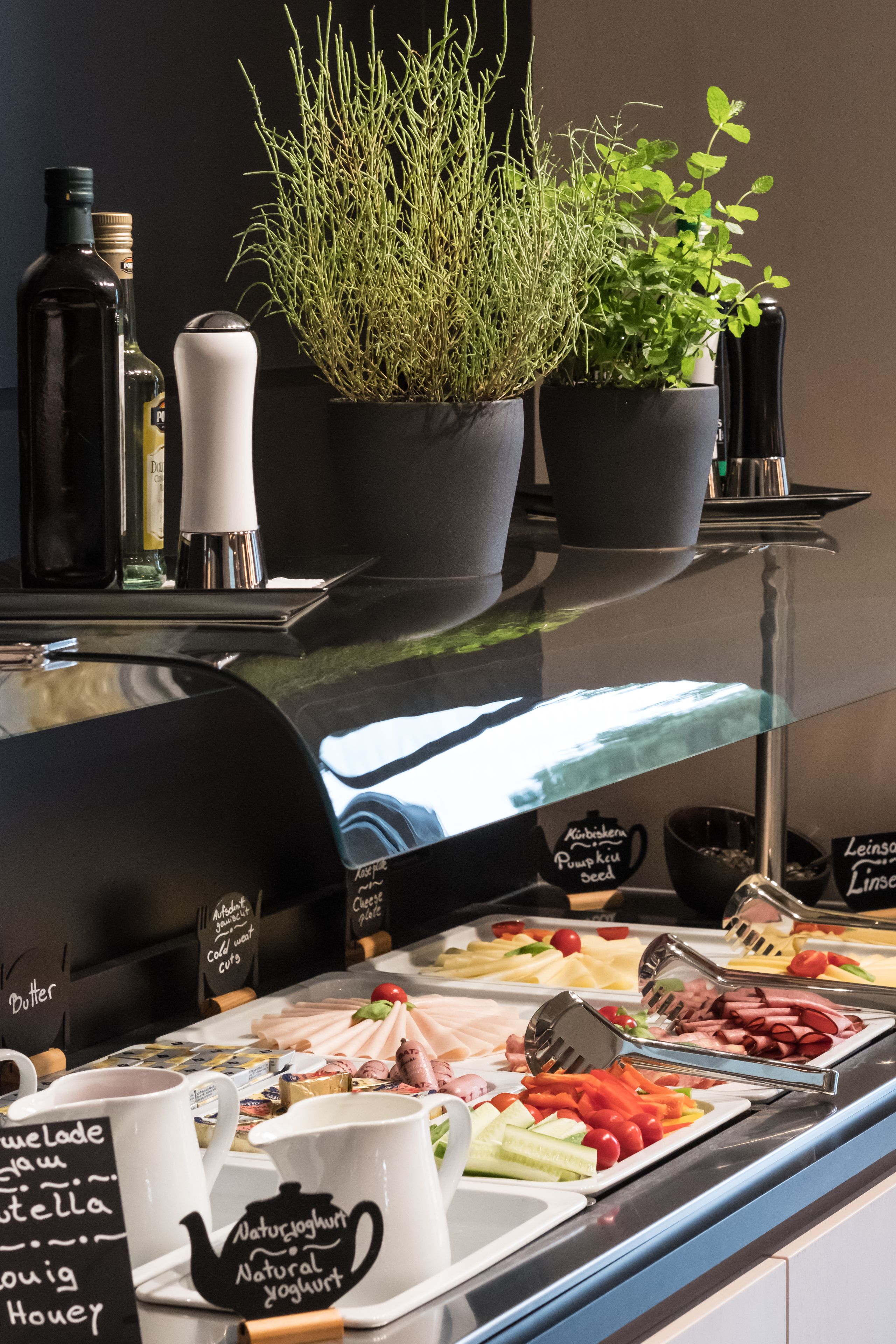 Een buffetgedeelte met diverse etenswaren, waaronder vlees, kaas en groenten. Er staan ook verschillende potplanten en flessen met sausjes op de toonbank.