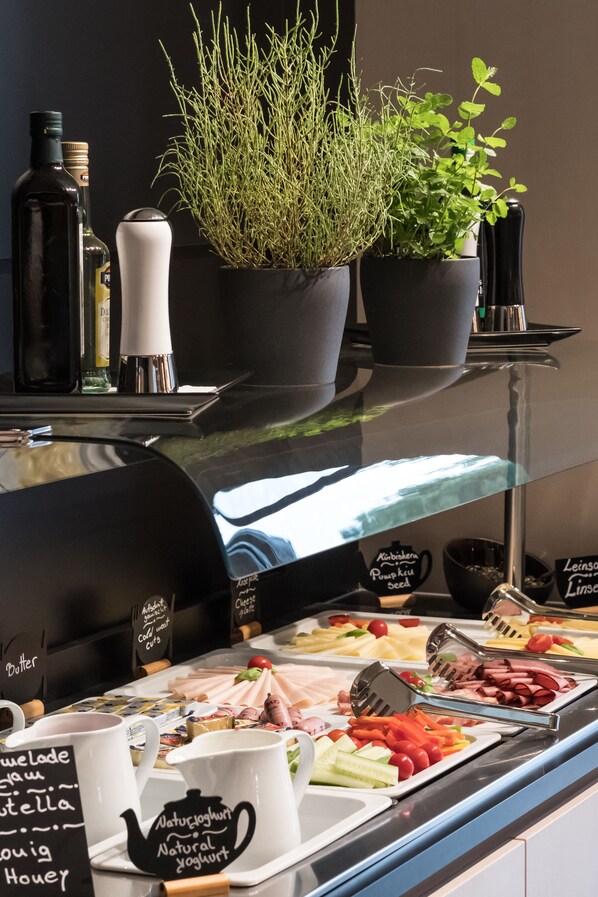A buffet area with various food items, including meats, cheeses, and vegetables. There are also several potted plants and condiment bottles on the counter. - Airbase Hotel (Kalsdorf bei Graz)