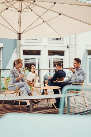 A group of people seated at an outdoor table with a large umbrella, surrounded by wooden chairs and a wooden deck. - Airbase Hotel (Kalsdorf bei Graz)