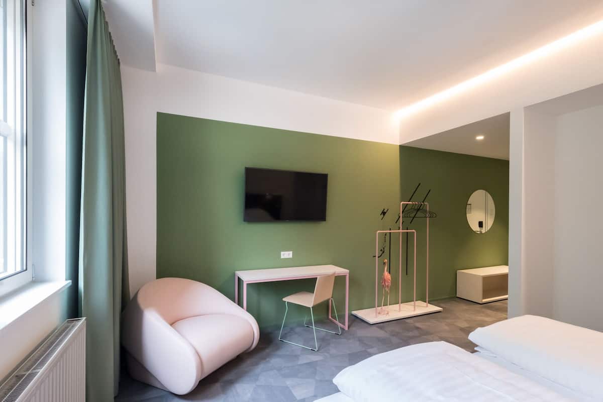 Una habitación de hotel moderna con una pared de acento verde, un televisor de pantalla plana, un escritorio con silla, una cama y un vestidor.