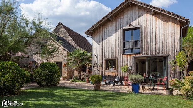 A rustic wooden house with a stone building in the background, a balcony, and outdoor seating.