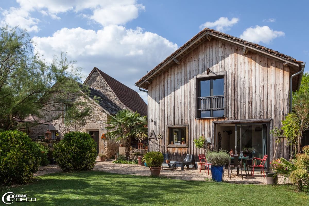 Une maison rustique en bois, avec un bâtiment en pierre en arrière-plan, un balcon et des sièges à l’extérieur.