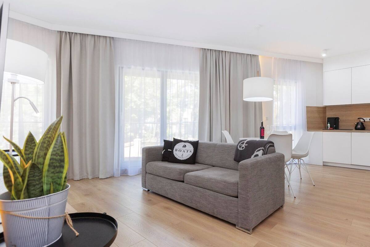 Ein modernes Wohnzimmer mit grauem Sofa, einem Esstisch mit Stühlen, einer Küchenzeile mit weißen Schränken und einem großen Fenster mit transparenten Vorhängen.