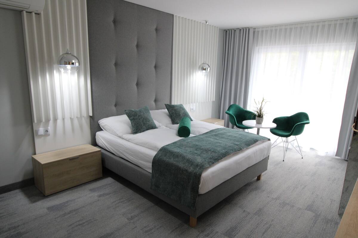 Ein modernes Hotelzimmer mit einem großen Bett, einem Nachttisch, einem kleinen runden Tisch, zwei grünen Stühlen und einer Topfpflanze.