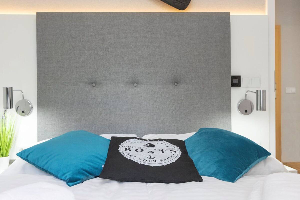 Ein modernes Hotelzimmer mit grauem Stoffbettkopfs, zwei blauen Kissen und einem schwarzen Kissen mit einem nautischen Motiv.