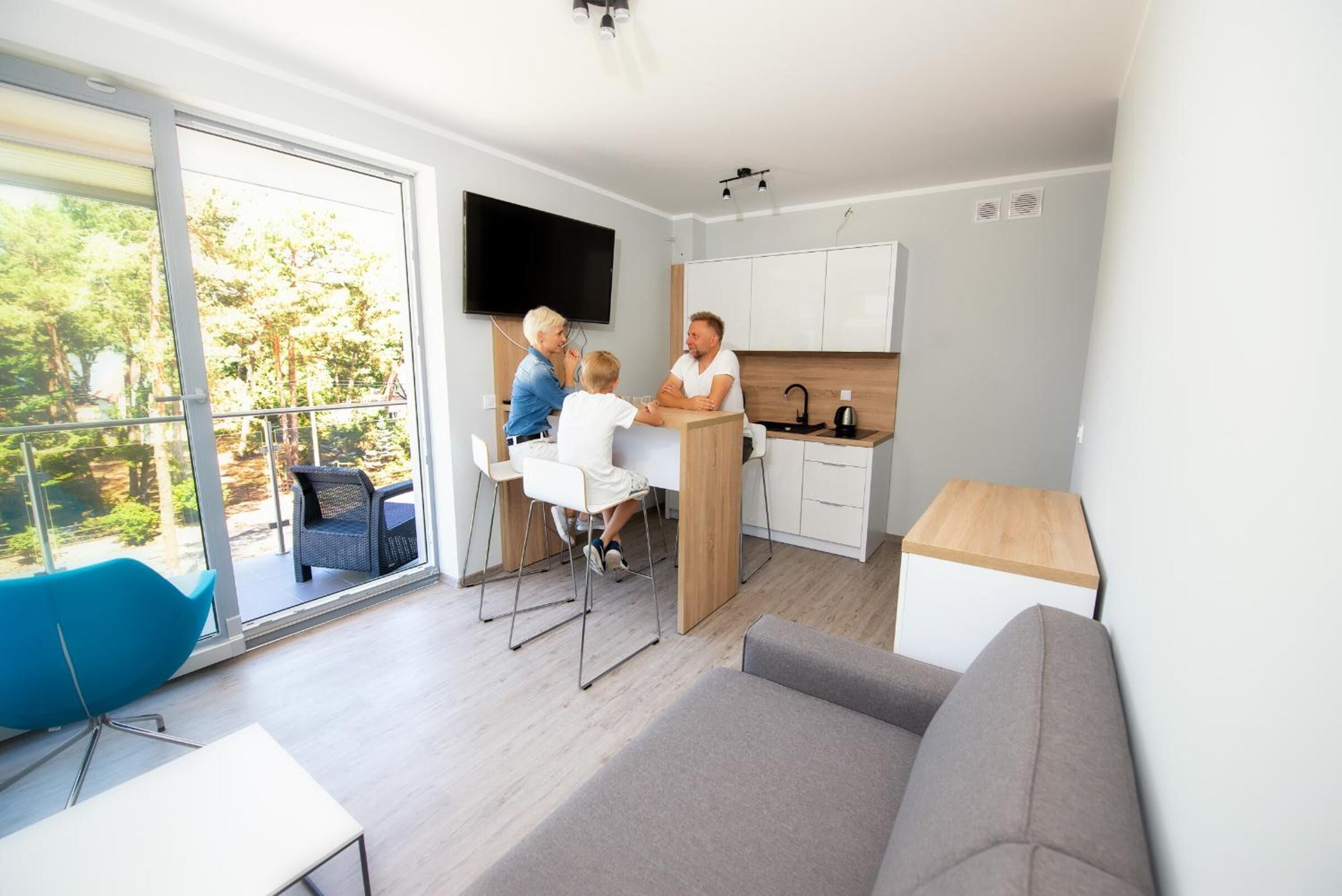 A modern living room with a sofa, a dining area with a table and chairs, a kitchen with cabinets and a TV, and a balcony with a chair.
