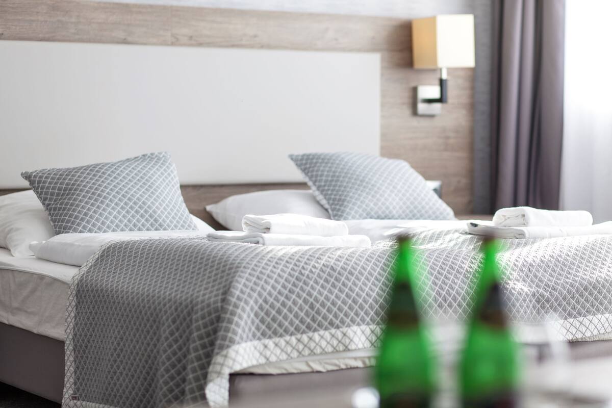Ein ordentlich bezogenes Bett mit grau gemustertem Bettbezug und Kissen, ein hölzernes Kopfteil und eine wandmontierte Leuchte.