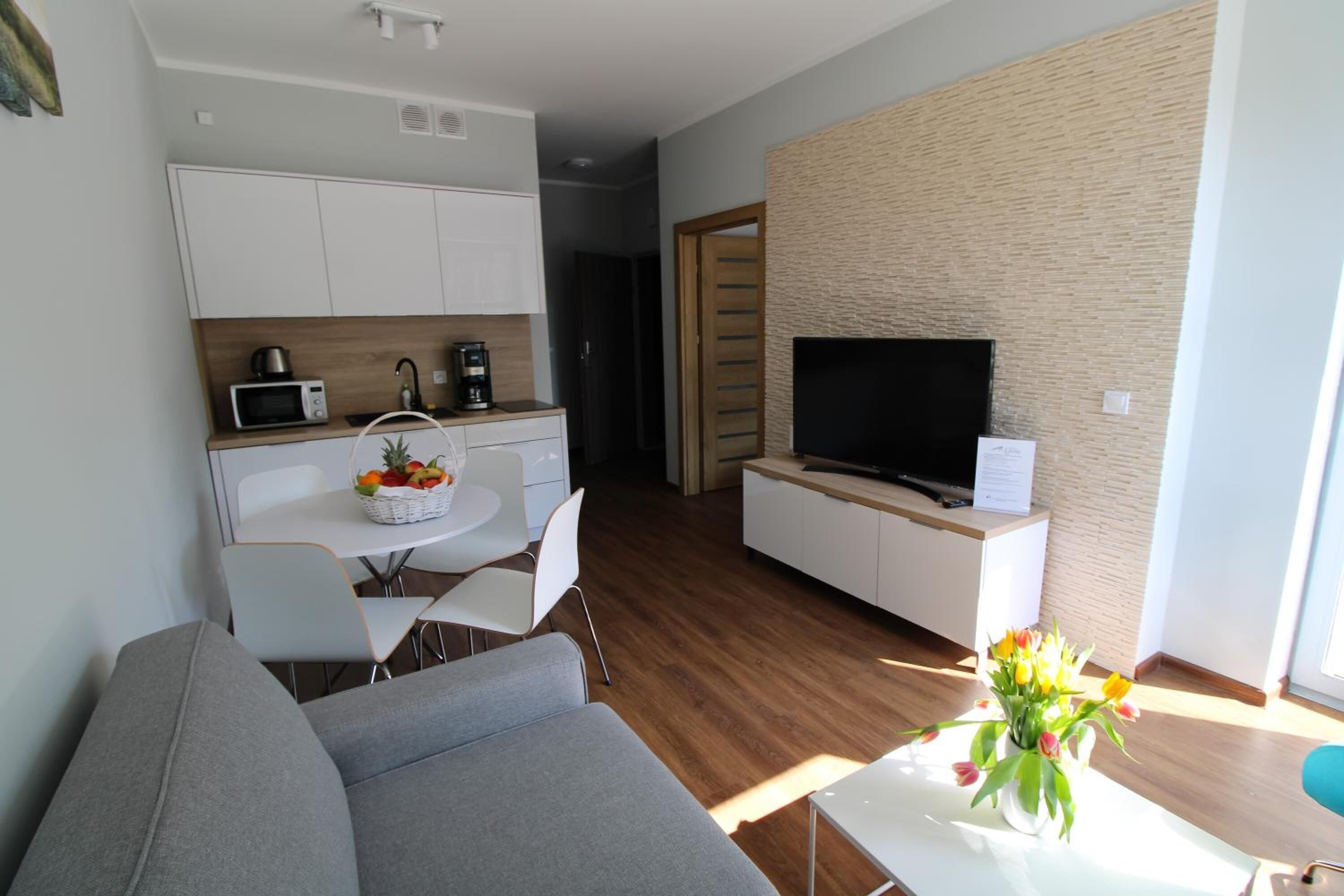 A modern living room with a grey sofa, a white coffee table with a vase of flowers, a flat-screen TV on a white cabinet, and a kitchen area with white cabinets and a microwave.