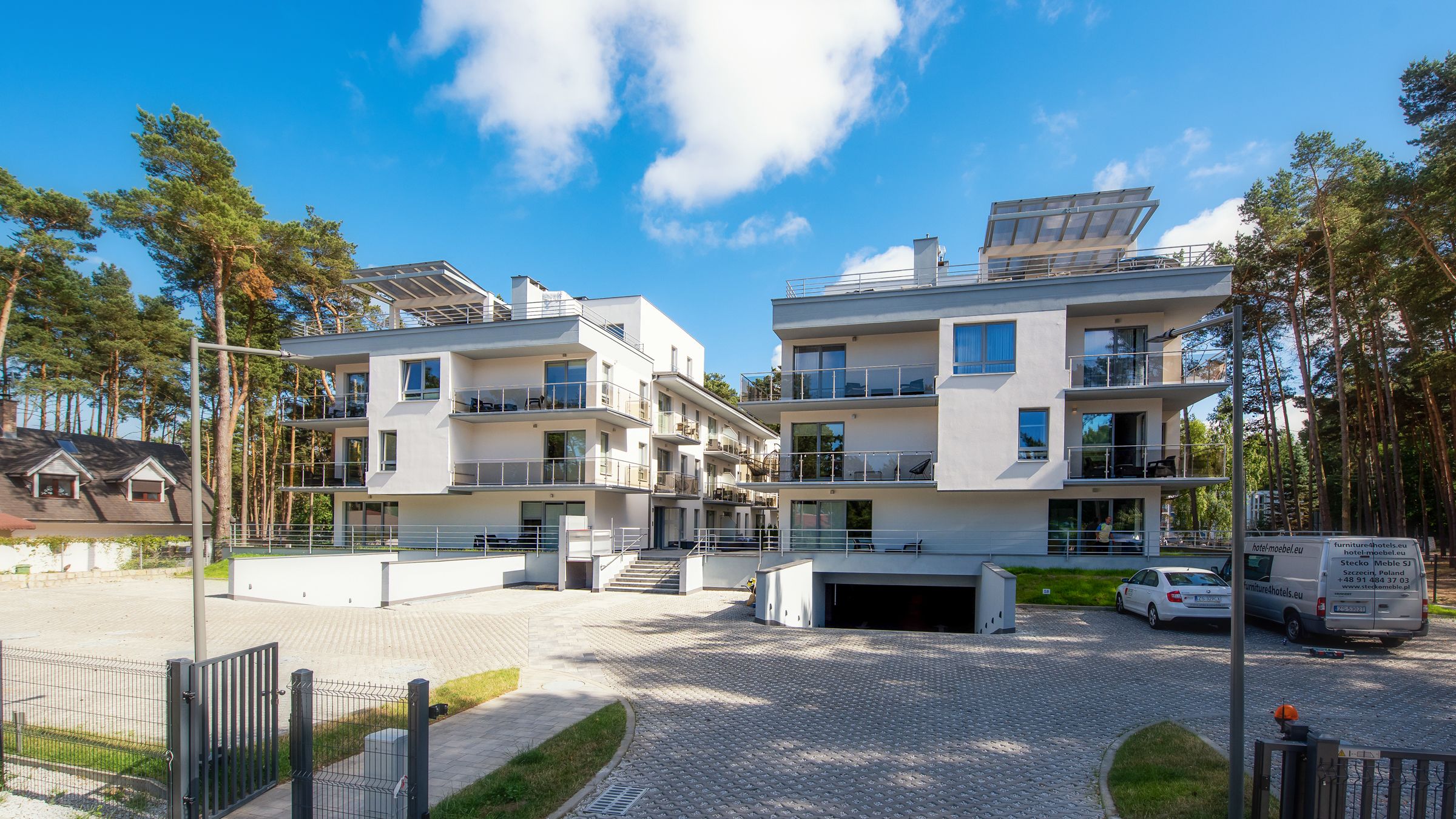 Un hotel moderno de varios pisos con balcones y paneles solares, rodeado de árboles y un camino pavimentado.