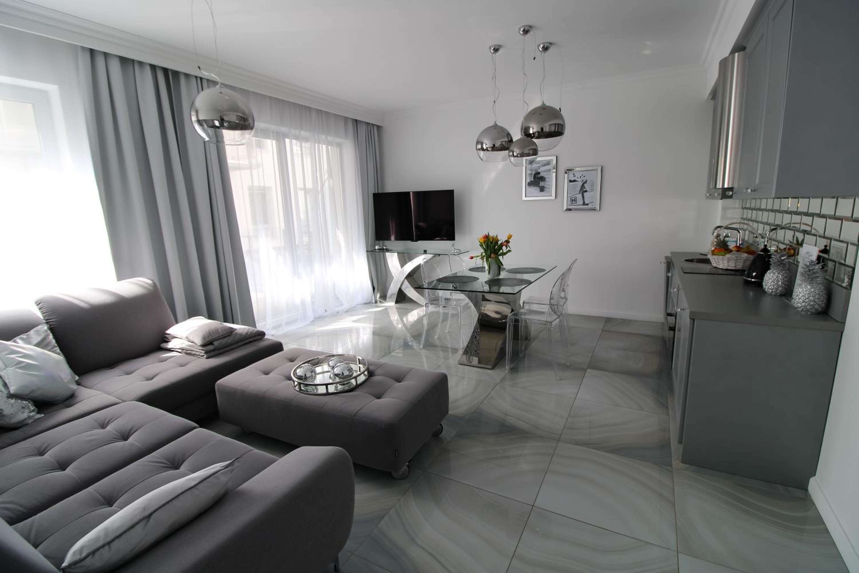 Una sala de estar moderna con un sofá gris, un reposapiés y una mesa de centro de vidrio. Hay una zona de comedor con una mesa y sillas de vidrio, y una cocina con armarios grises y un salpicadero de azulejos.